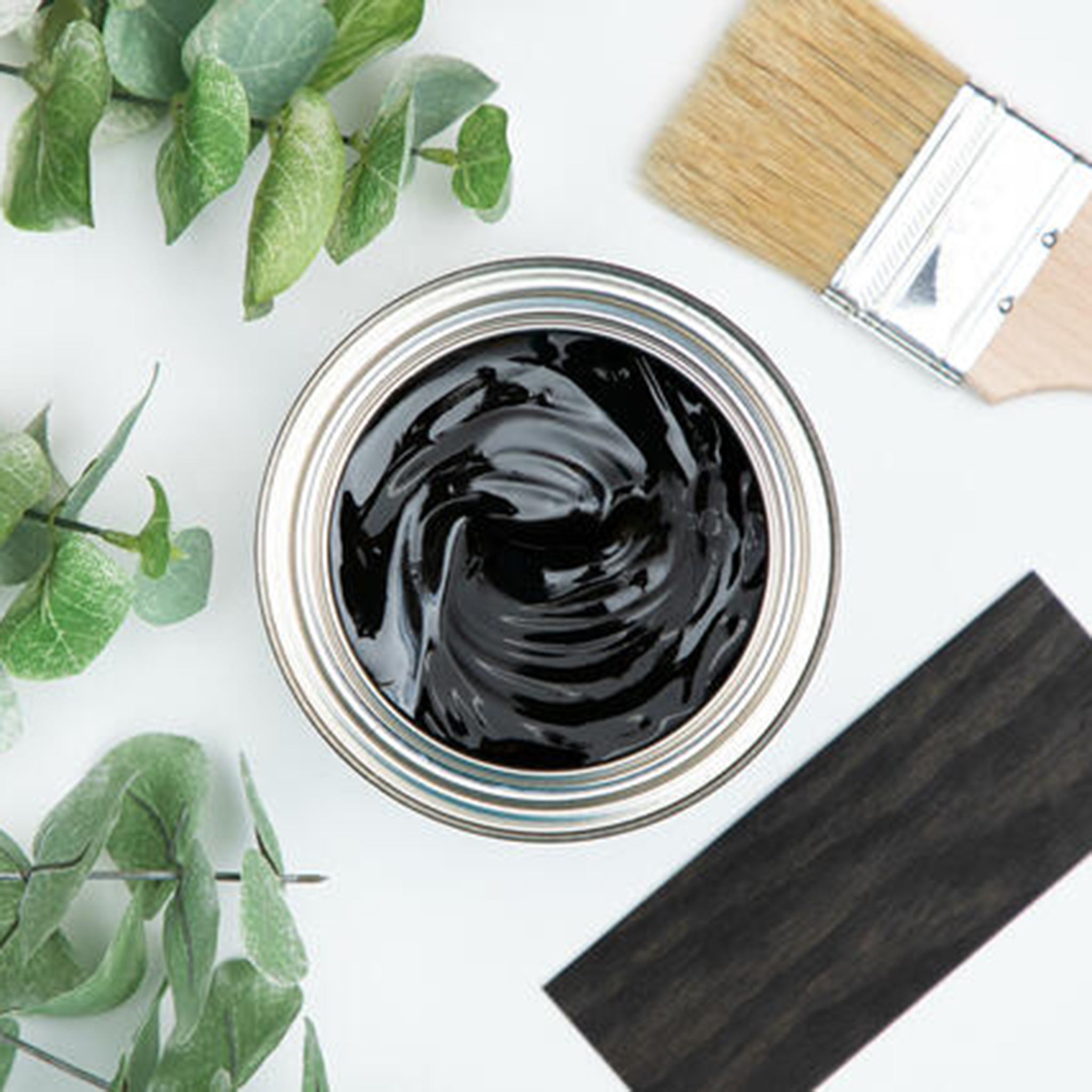 A arial view of an open can of Dixie Belle's No Pain Gel Stain in Colonial Black is against a white background. The can is surrounded by a paint brush, green foliage, and a wood swatch featuring the stain color.