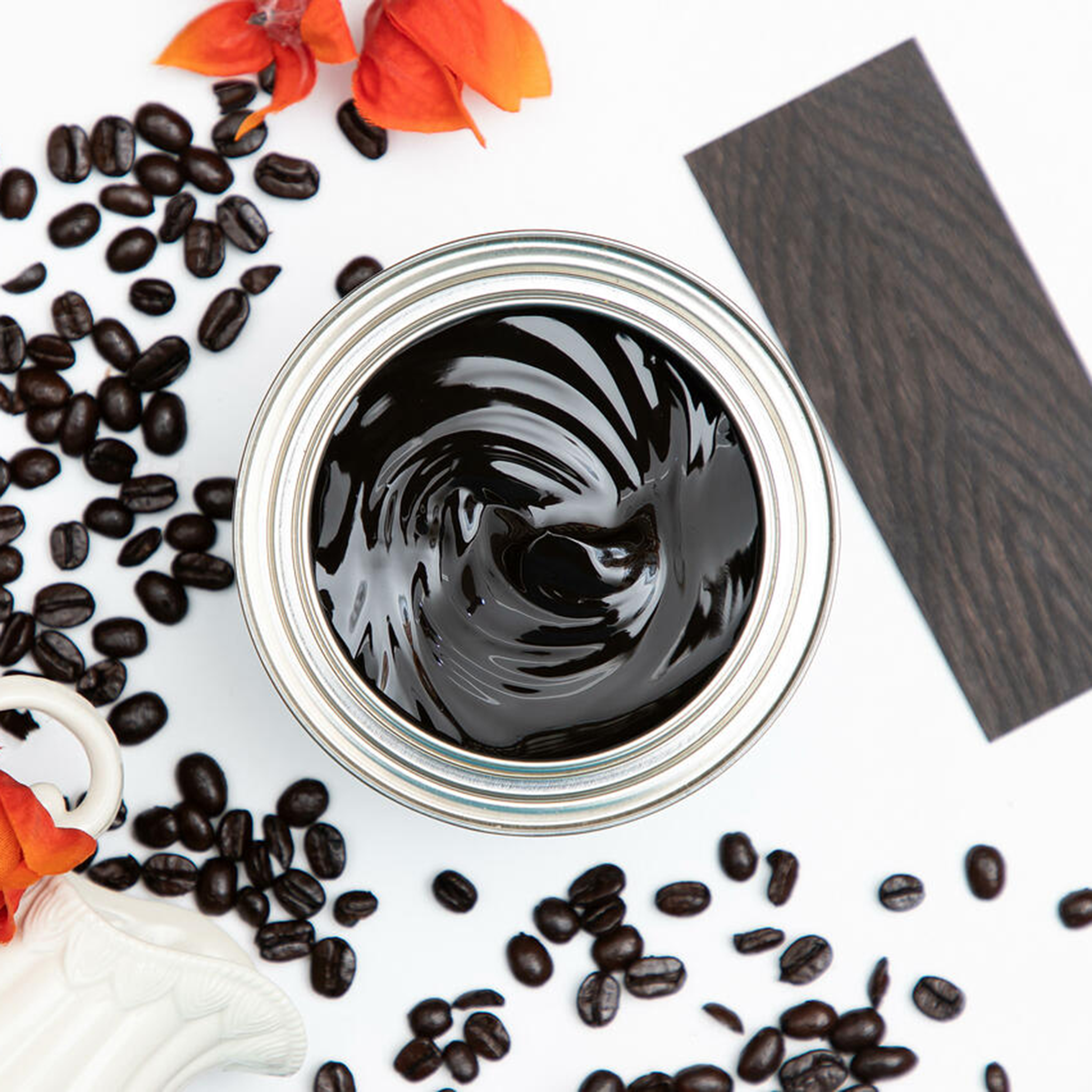 A arial view of an open can of Dixie Belle's No Pain Gel Stain in Espresso is against a white background. The can is surrounded by espresso beans, red-orange flowers, and a wood swatch featuring the stain color.