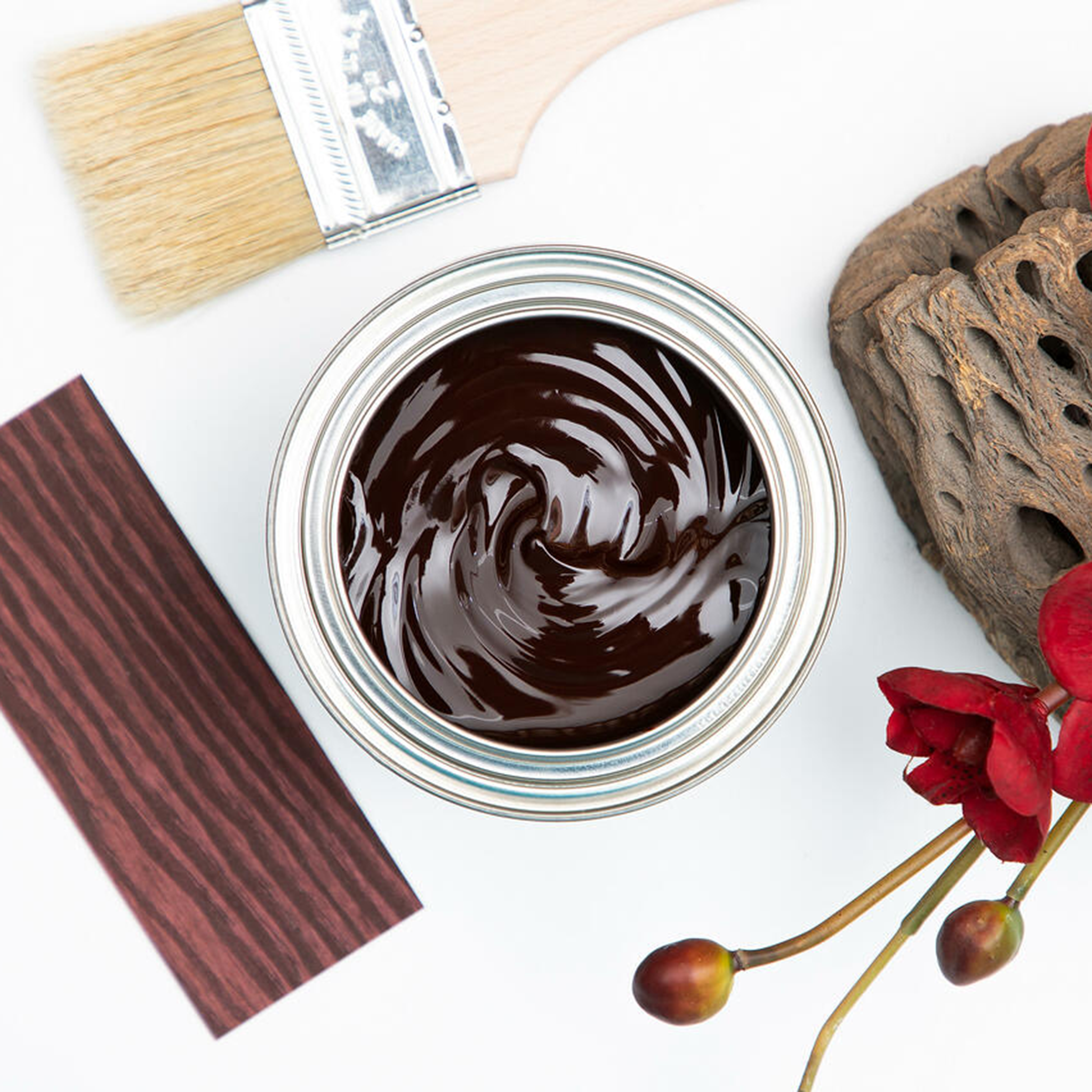 A arial view of an open can of Dixie Belle's No Pain Gel Stain in Georgian Cherry is against a white background. The can is surrounded by a paint brush, a piece of driftwood with red flowers, and a wood swatch featuring the stain color.