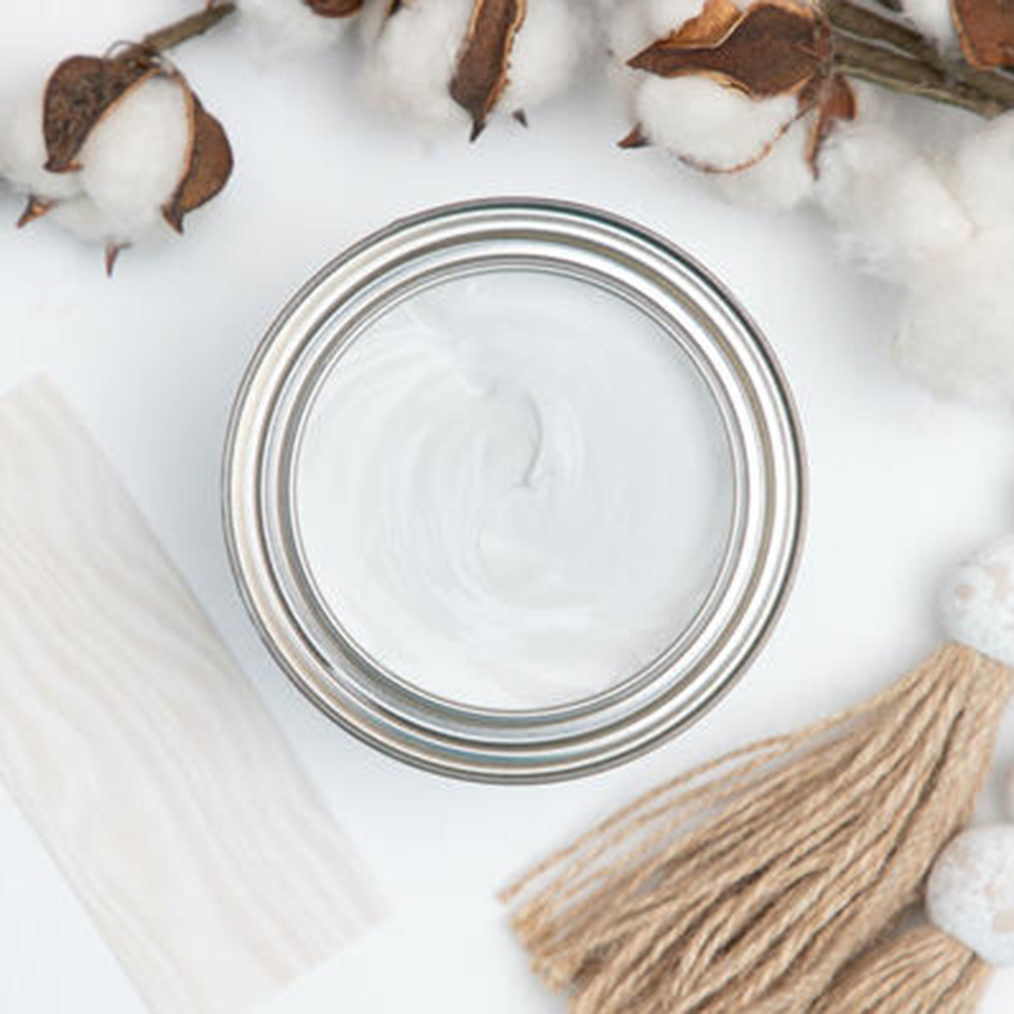 A arial view of an open can of Dixie Belle's No Pain Gel Stain in Picklin White is against a white background. The can is surrounded by twine tassles, real cotton, and a wood swatch featuring the stain color.