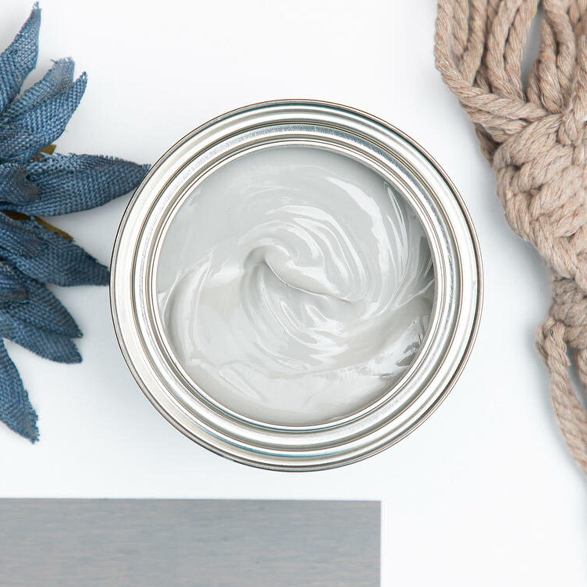 A arial view of an open can of Dixie Belle's No Pain Gel Stain in Weathered Gray is against a white background. The can is surrounded by rope, a cloth blue flower, and a wood swatch featuring the stain color.