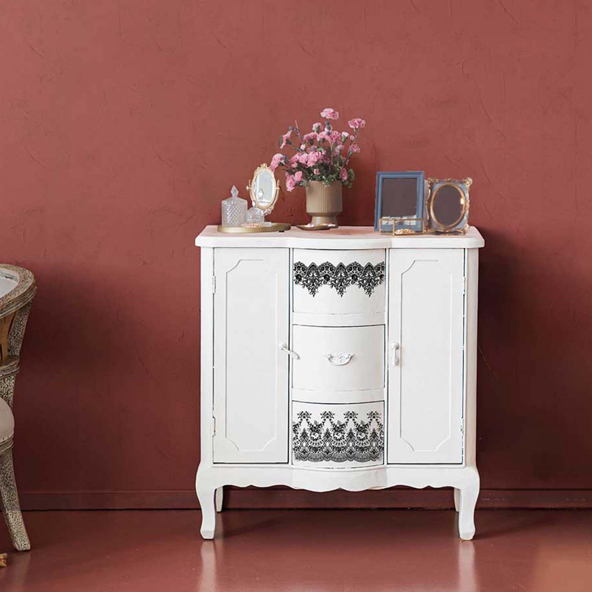 A tall white vintage cabinet with black lace design across the 2 drawers. Vintage vanity set, frames and vase are displayed on top.