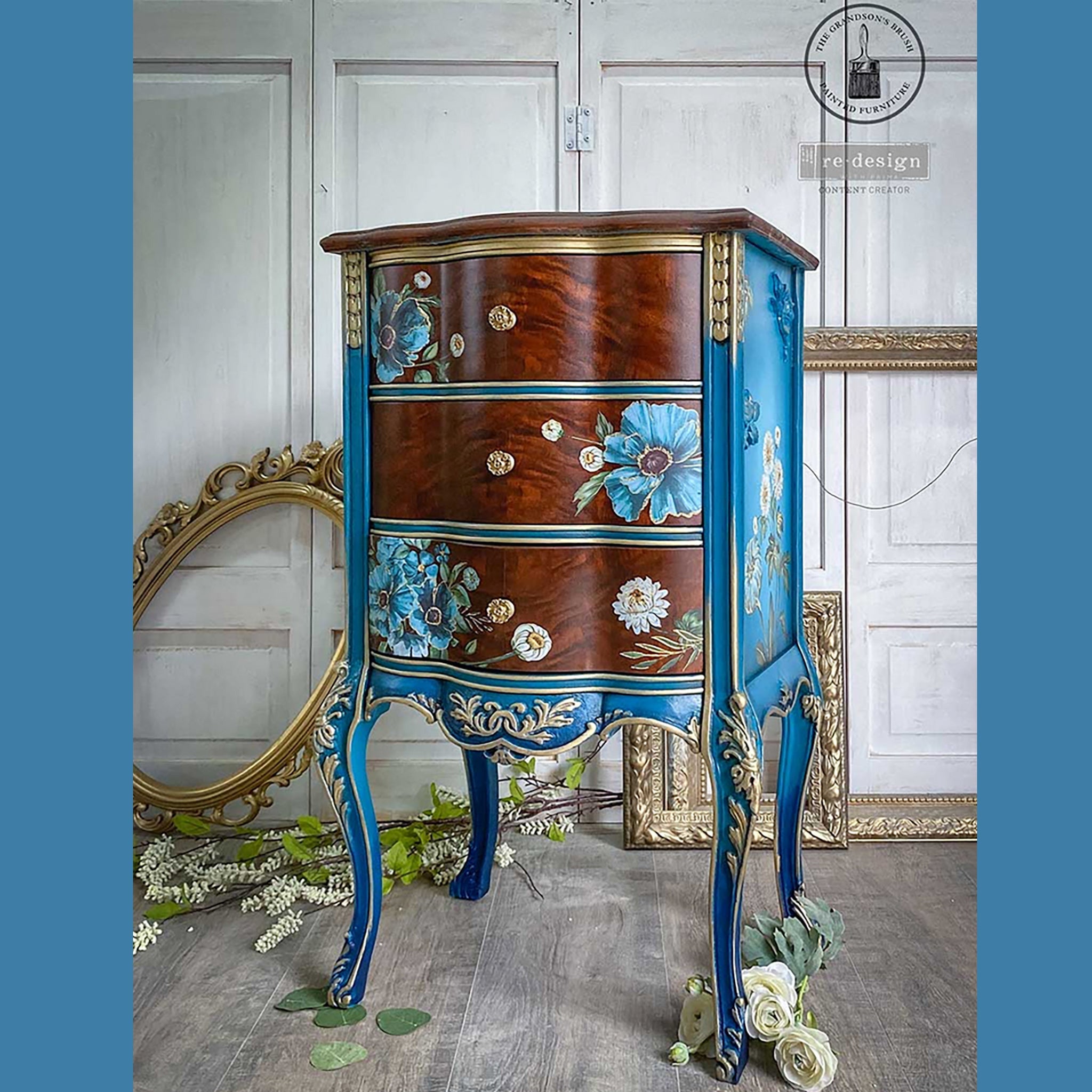 A small 3-drawer nightstand refurbished by The Grandson's Brush is painted blue with gold accents and features Gilded Floral on the sides and on its natural wood stained drawers.