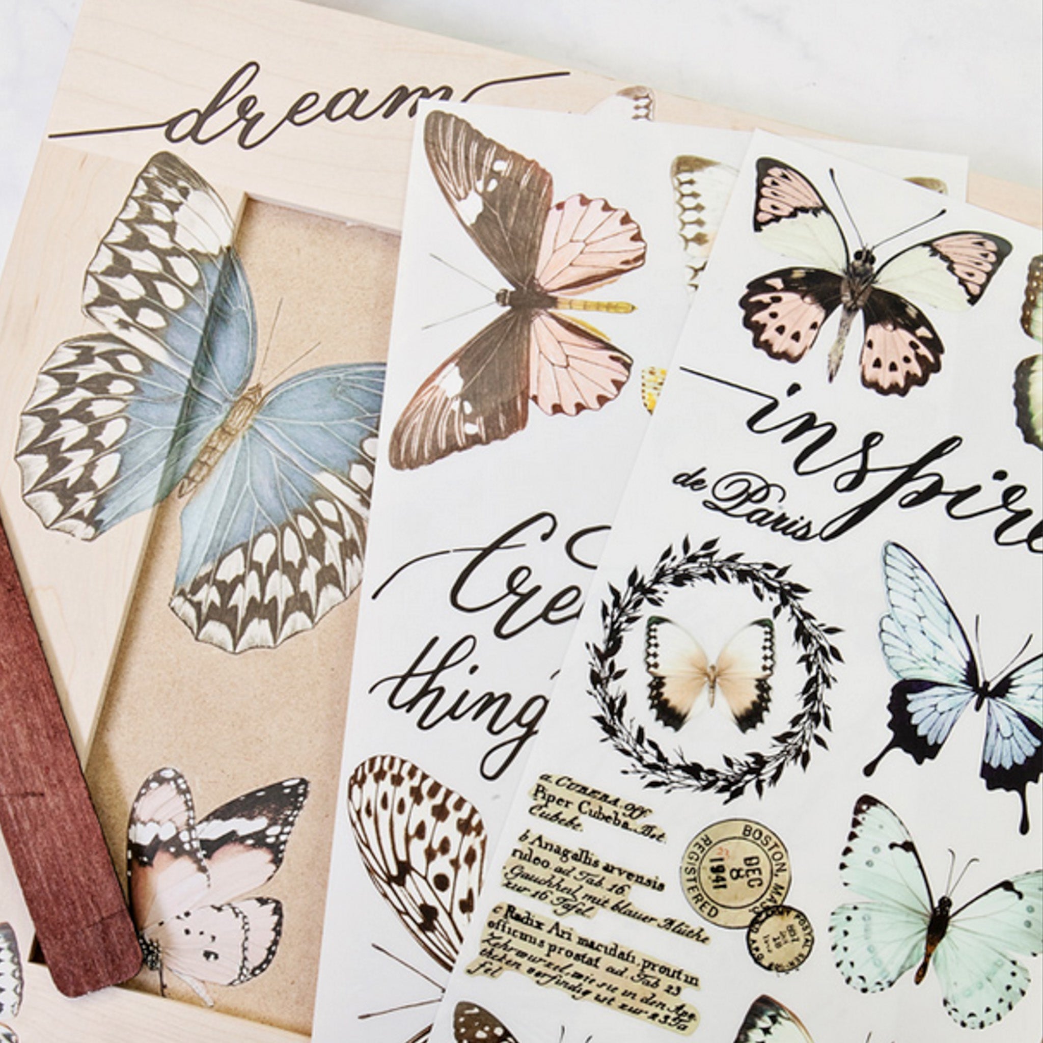 A natural wood photo frame craft features ReDesign with Prima's Papillon Collection small transfer on it. Two sheets of the transfer sit on the project.