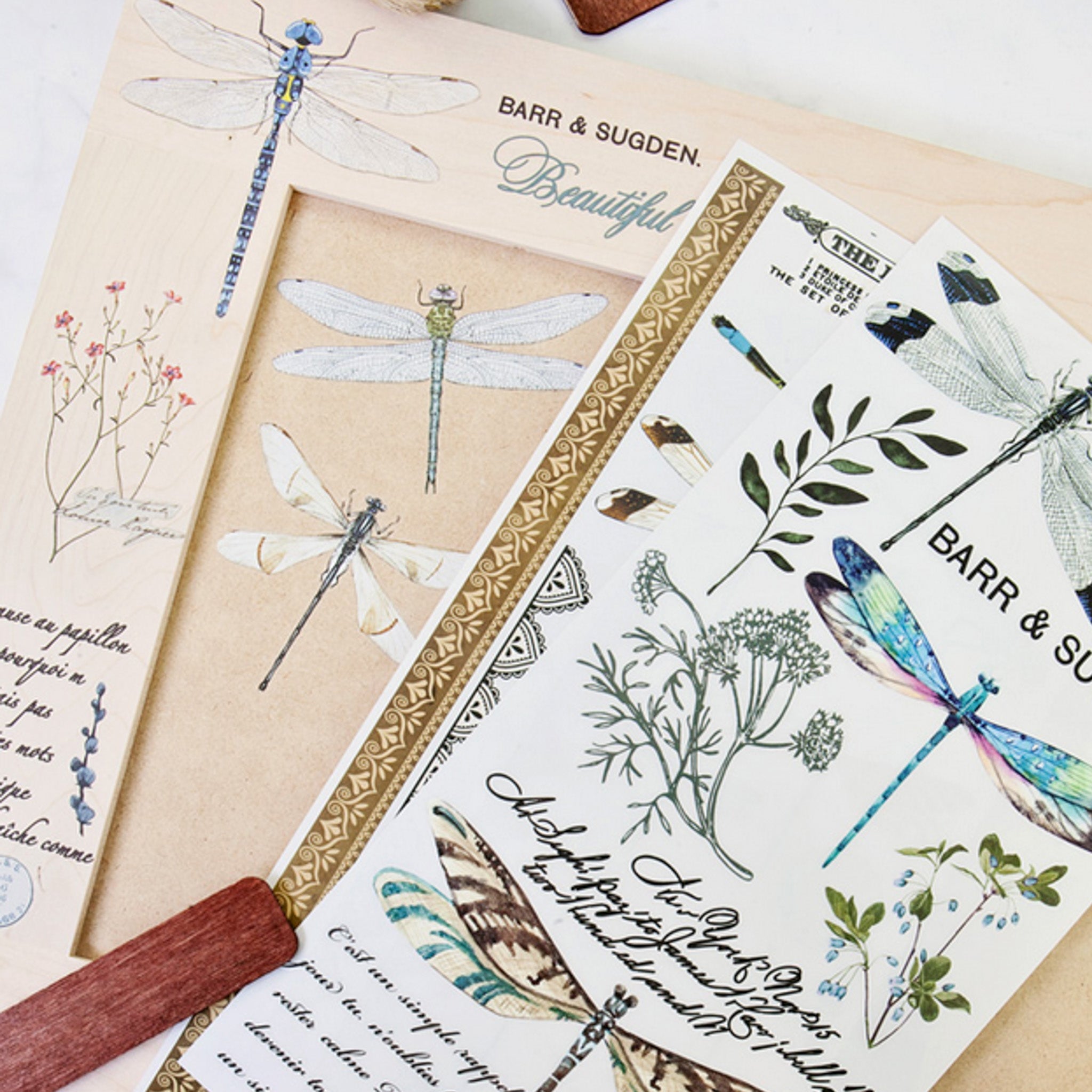 A small wood photo project features ReDesign with Prima's Spring Dragonfly small transfer on it. Two sheets of the transfers sit on the craft project.