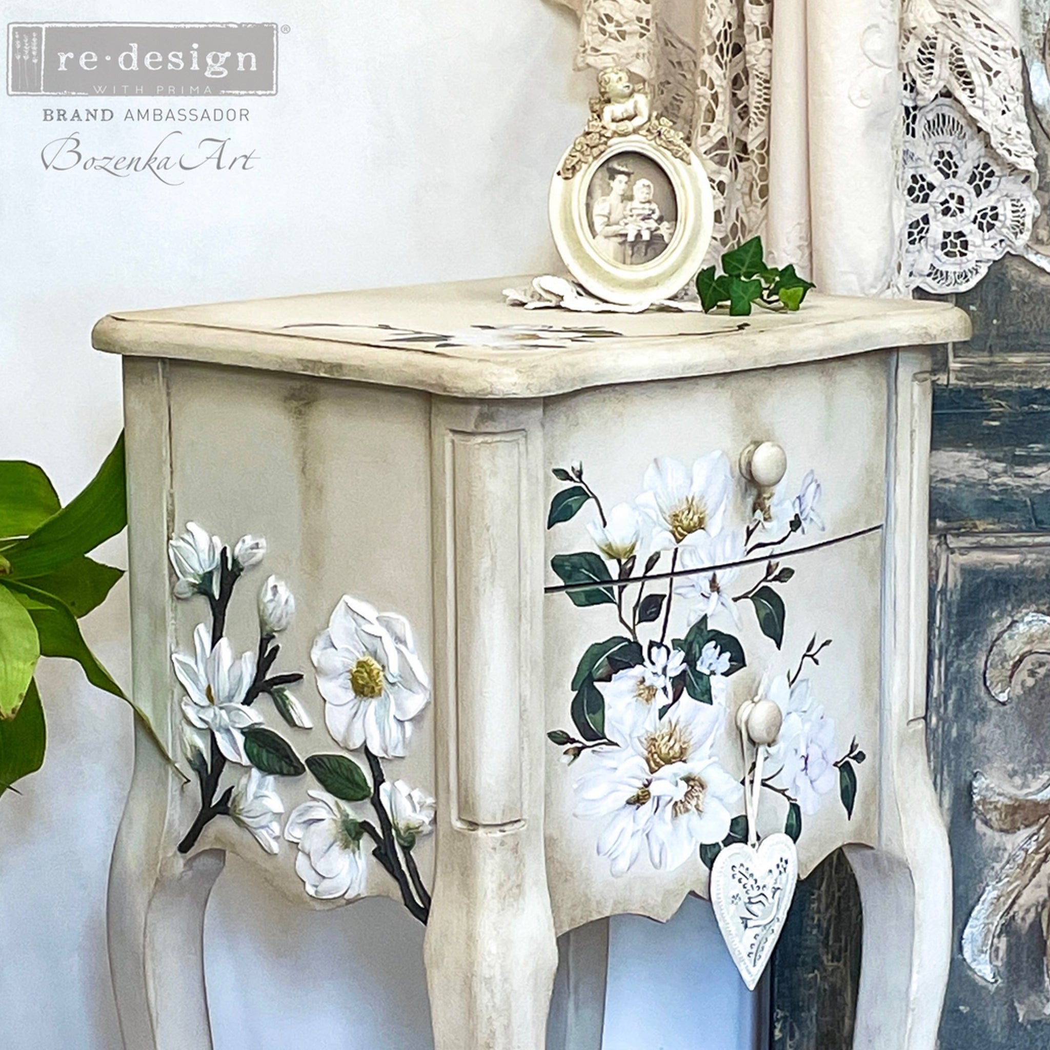 A close-up of a vintage, small, 2-drawer nightstand refurbished by Bozenka Art is painted antique white and features ReDesign with Prima's White Magnolia Small transfer on it.