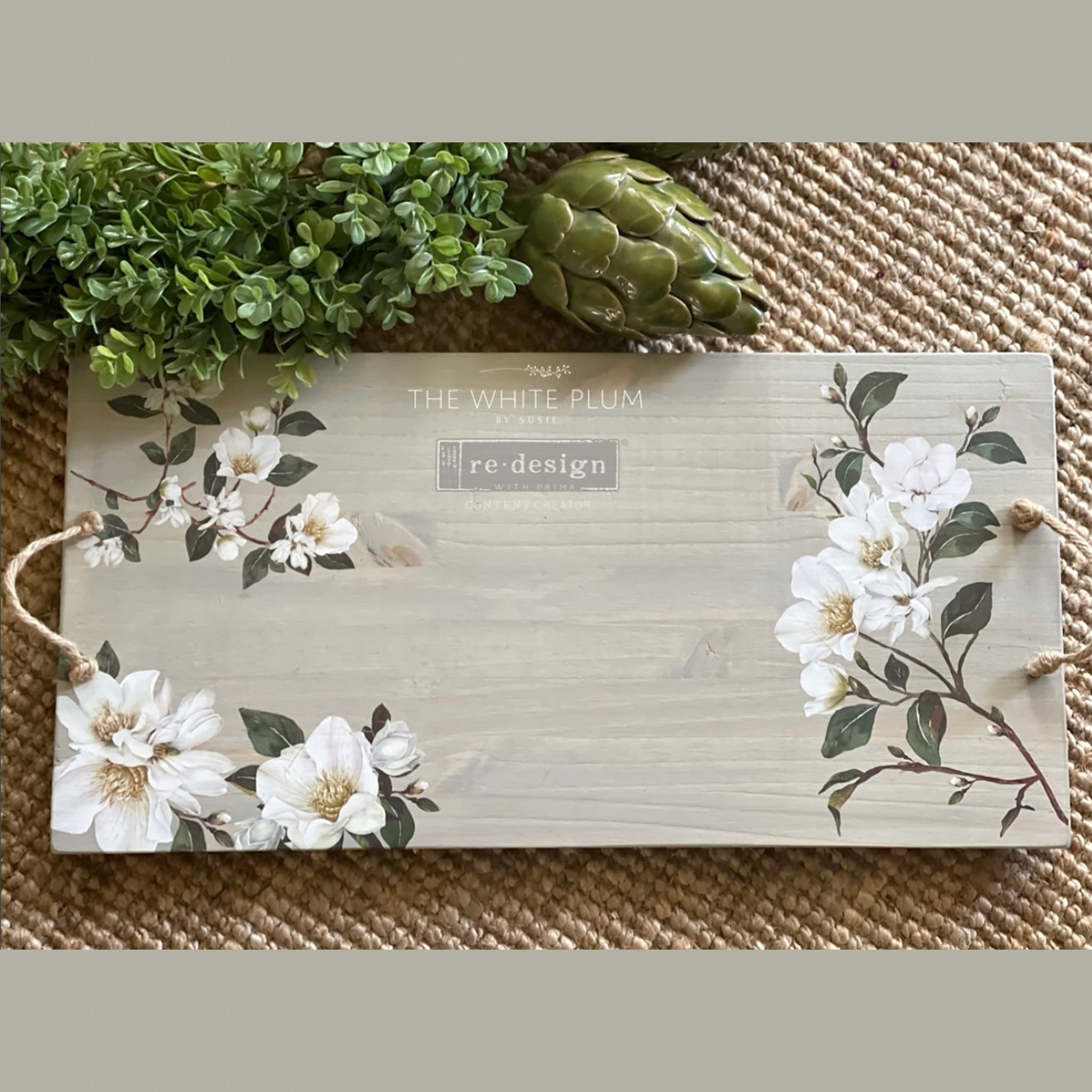 A wood serving tray with a rope handle is white-washed and features ReDesign with Prima's White Magnolia small transfer on it.