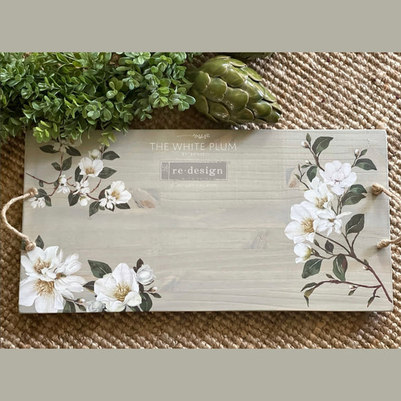 A wood serving tray with a rope handle is white-washed and features ReDesign with Prima's White Magnolia small transfer on it.