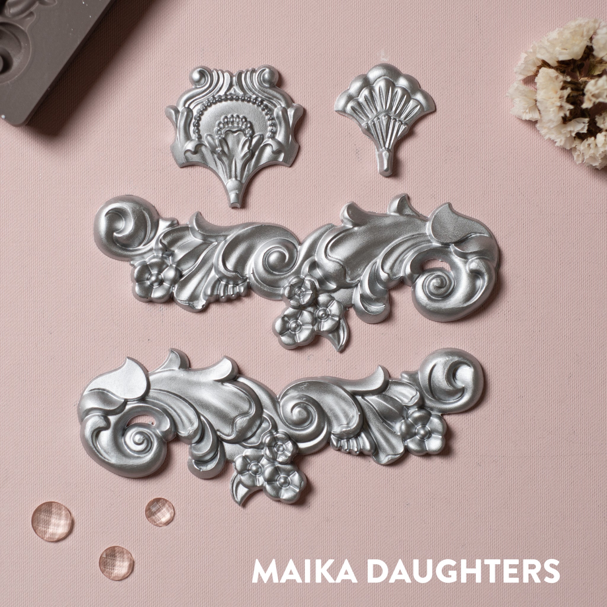Groeneville Crest silver mold castings on a pink background. A white Maika Daughters logo on the bottom right.