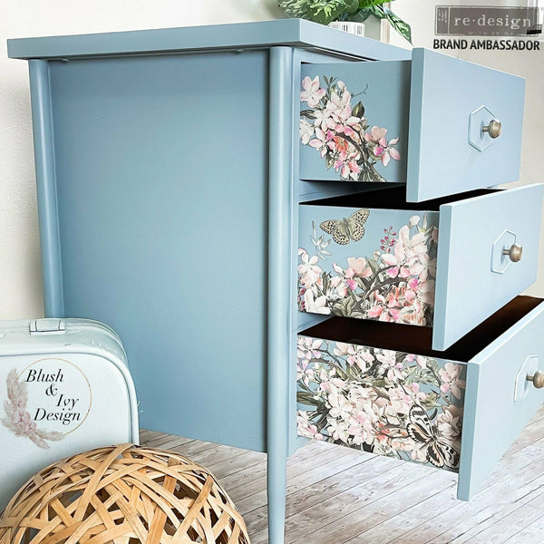 A blue dresser with the Blossom Botanical transfer on the sides of each drawer. A Blush & Ivy Design logo in the bottom left corner. A Redesign with Prima Brand Ambassador logo in the top right.