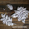 Wooden background with silver castings of the Perfect Poinsettia mold. A white Maika Daughters logo is in the bottom right corner.