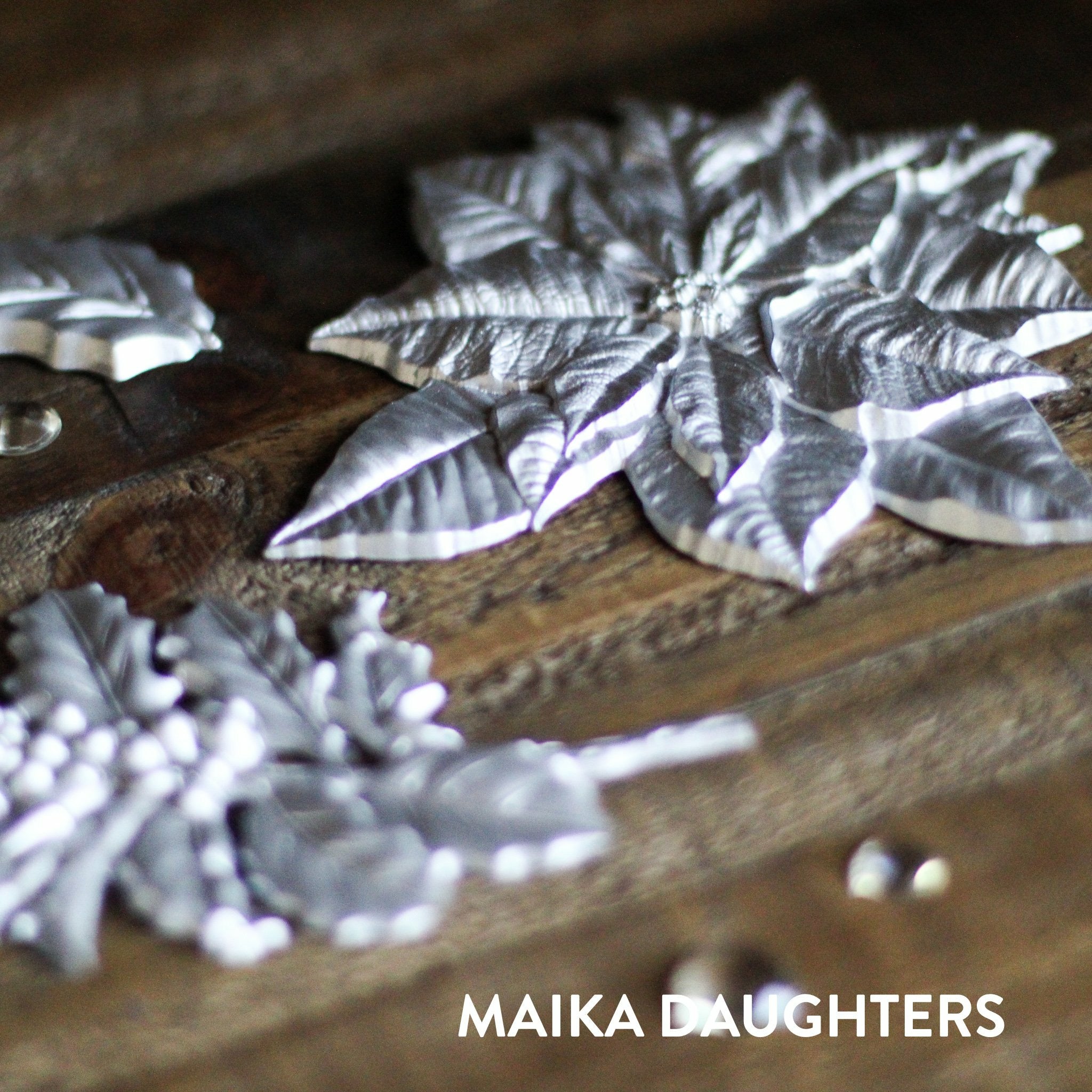 Wooden background with a angled view of silver castings of the Perfect Poinsettia mold. A white Maika Daughters logo is in the bottom right corner.