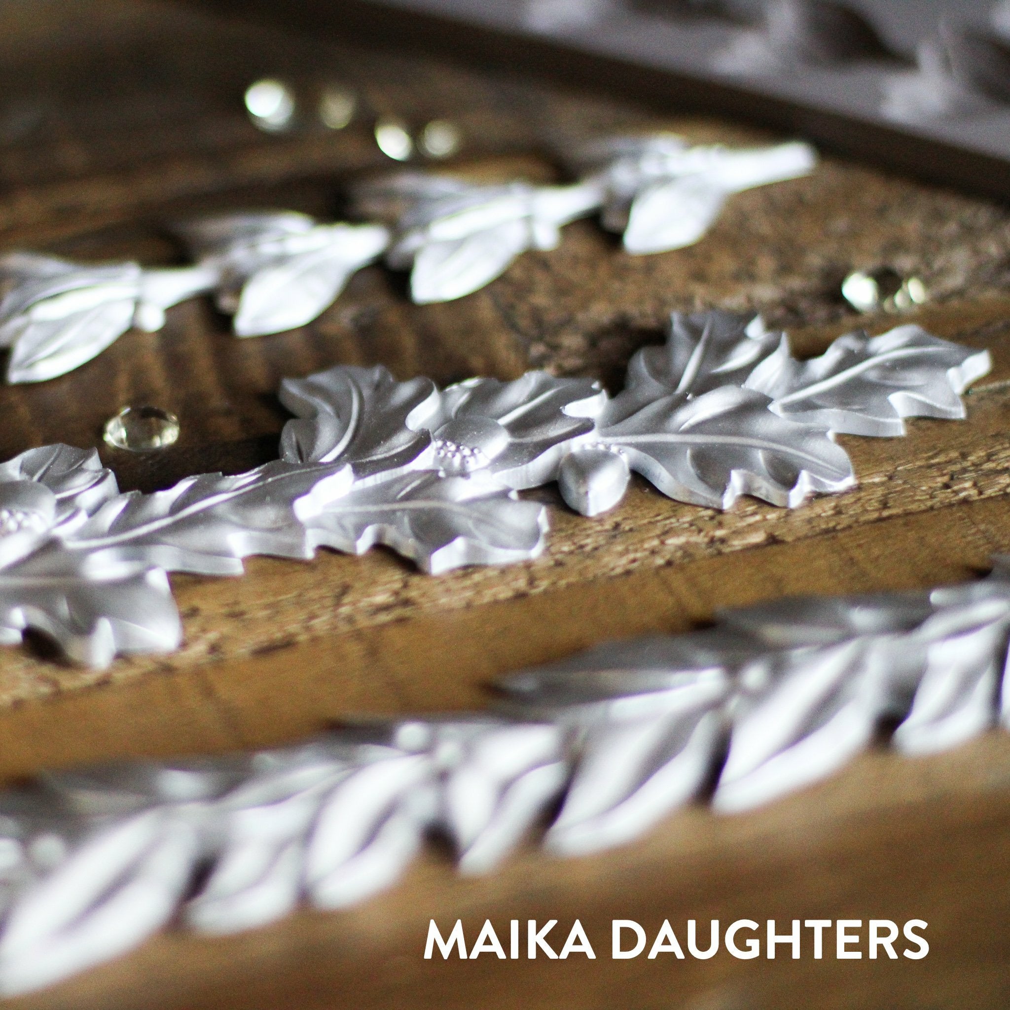 Wooden background with a angled view of silver castings of the Louelle Borders mold. A white Maika Daughters logo is in the bottom right.