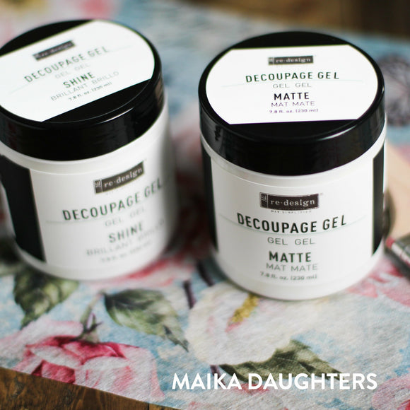 Two white containers with a black lid and a white logo. The container on the left reads: Redesign. Decoupage Gel. Gel. Gel. Shine. Brillant. Brillo. 7.8 fl. oz. (230ml) The container on the right reads: Redesign. Decoupage Gel. Gel. Gel. Matte. Mat. Mate. 7.8 fl. oz. (230ml) On the bottom right corner is white text reading Maika Daughters.