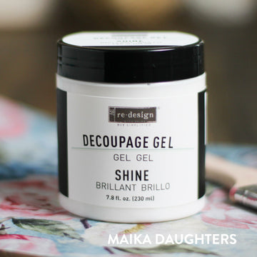 A white container with a black lid and a black text logo reading: Redesign diy simplified. Decoupage Gel. Gel. Gel. Shine. Brillant. Brillo. 7.8 fl. oz. (230 ml) On the bottom right corner is white text reading Maika Daughters.