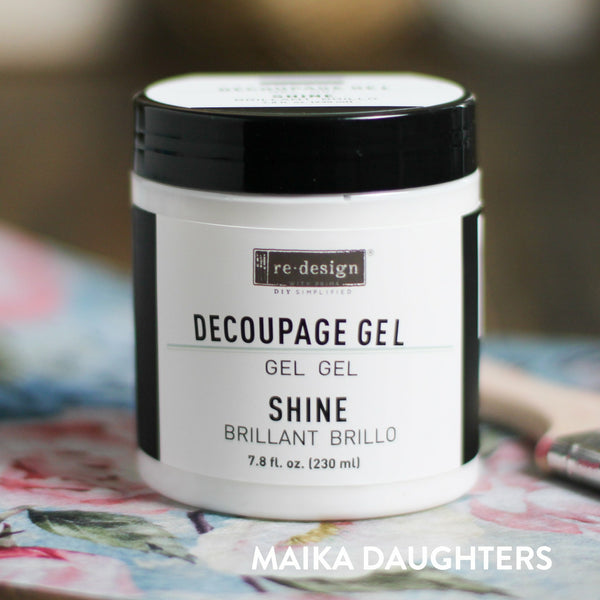 A white container with a black lid and a black text logo reading: Redesign diy simplified. Decoupage Gel. Gel. Gel. Shine. Brillant. Brillo. 7.8 fl. oz. (230 ml) On the bottom right corner is white text reading Maika Daughters.