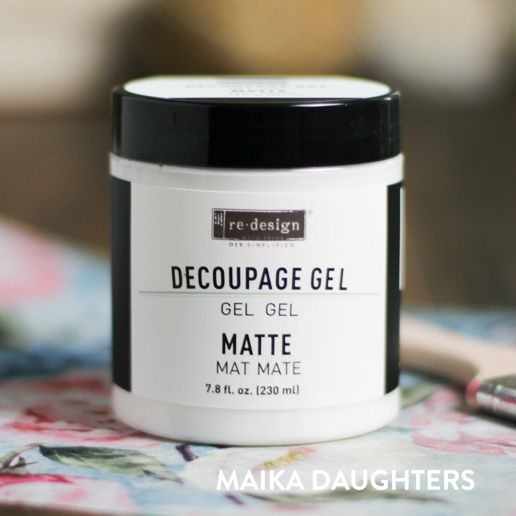 A white container with a black lid and a black text logo reading: Redesign diy simplified. Decoupage Gel. Gel. Gel. Matte. Mat. Mate. 7.8 fl. oz. (230 ml) On the bottom right corner is white text reading Maika Daughters.