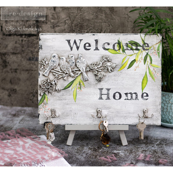 A small piece of wood turned into a key holder refurbished by Olga Kuvlavskaya, a ReDesign with Prima Content Creator, is painted white with text that reads: Welcome Home and features th Avian Love silicone mould on it.