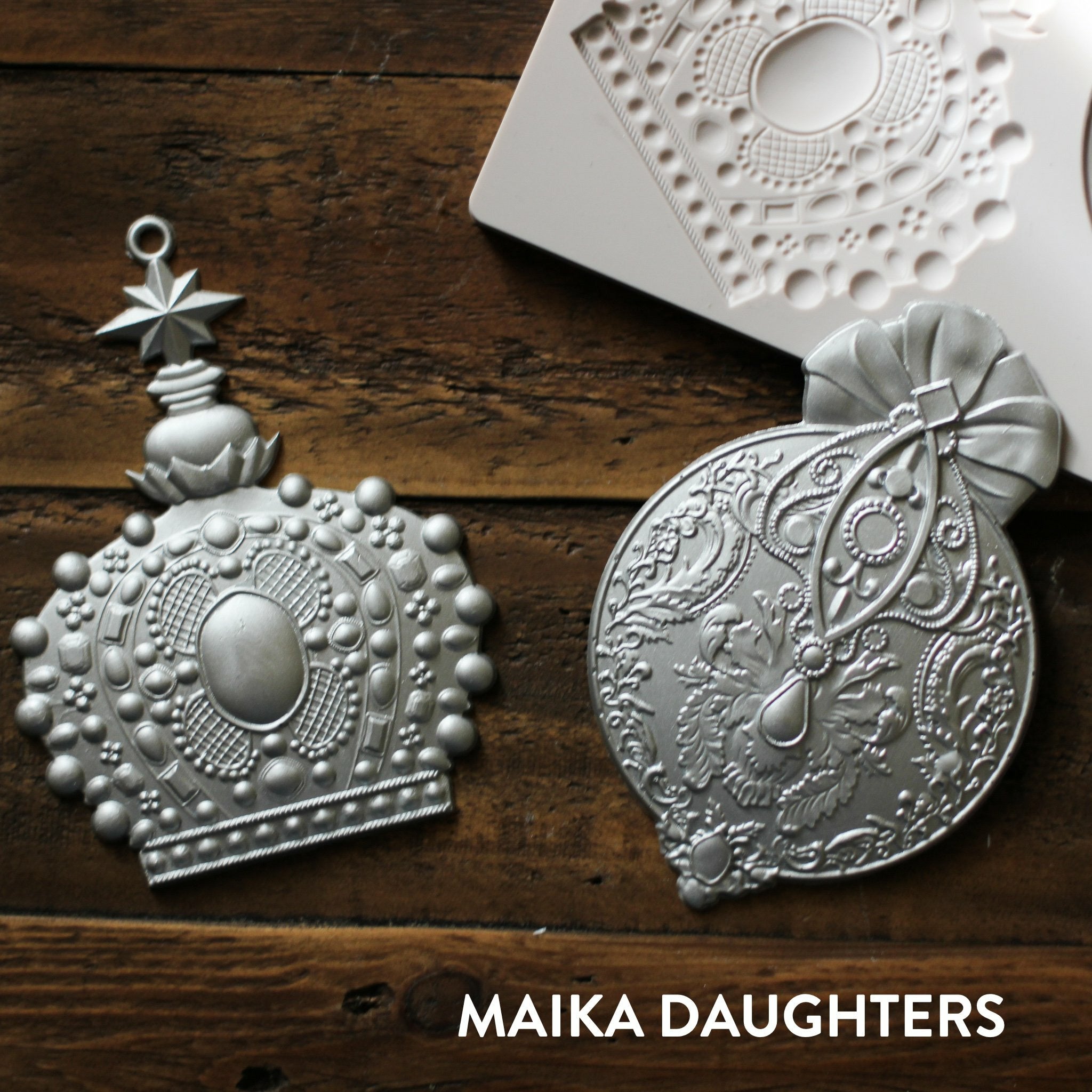 Wooden background with the victorian adornments castings painted in a metallic silver. A white Maika Daughters logo is sat in the bottom right corner.