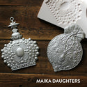 Wooden background with the victorian adornments castings painted in a metallic silver. A white Maika Daughters logo is sat in the bottom right corner.