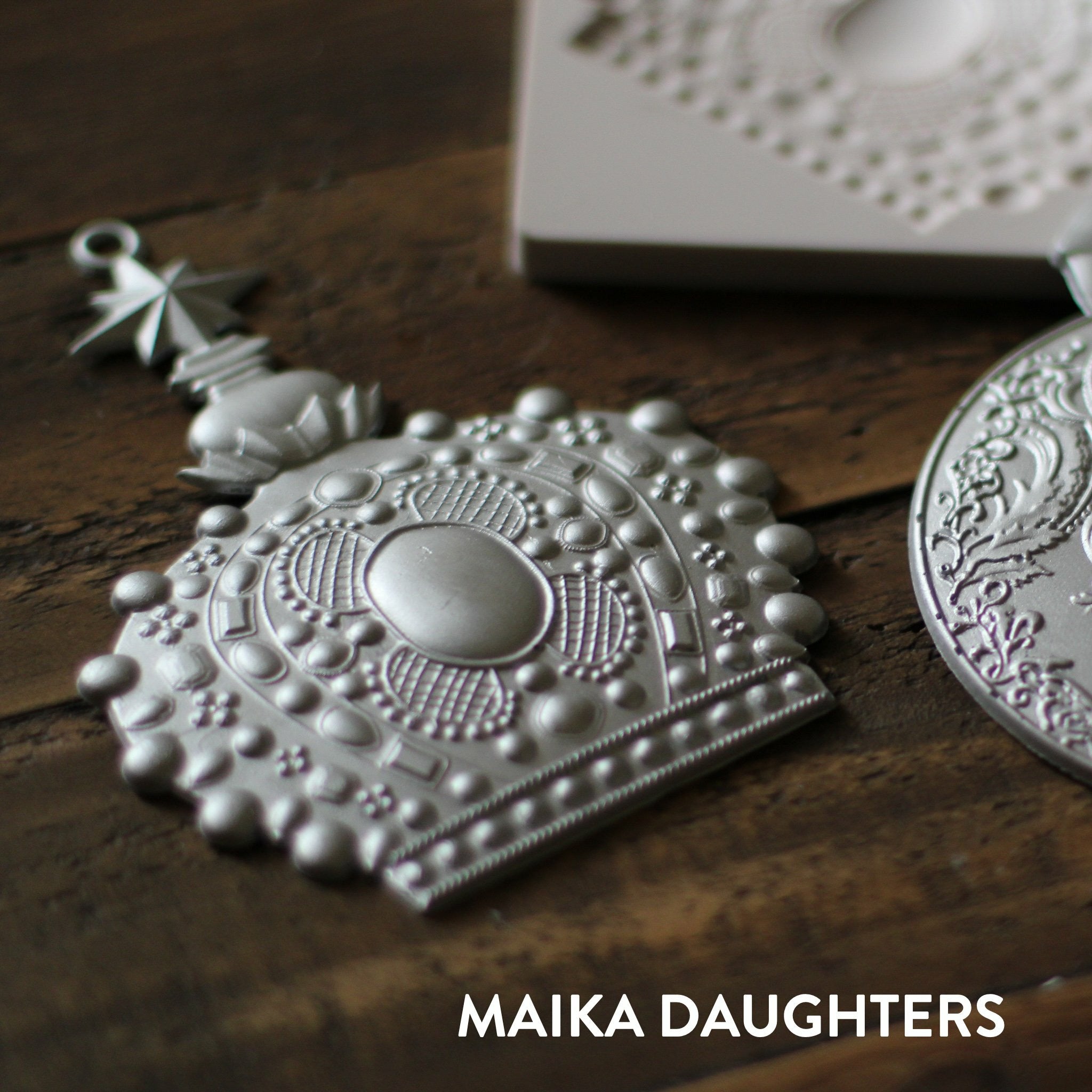 Wooden background with a angled view of the victorian adornments castings painted in a metallic silver. A white Maika Daughters logo is in the bottom right corner.