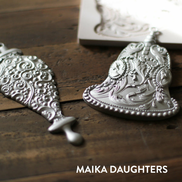 Wooden background with a angled view of two silver bell castings painted in a metallic silver color. A white maika daughters logo is in the bottom right corner.