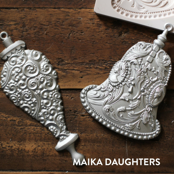 Wooden background with castings of the two silver bells mold painted in a metallic silver color. A white maika daughters logo is in the bottom right corner.