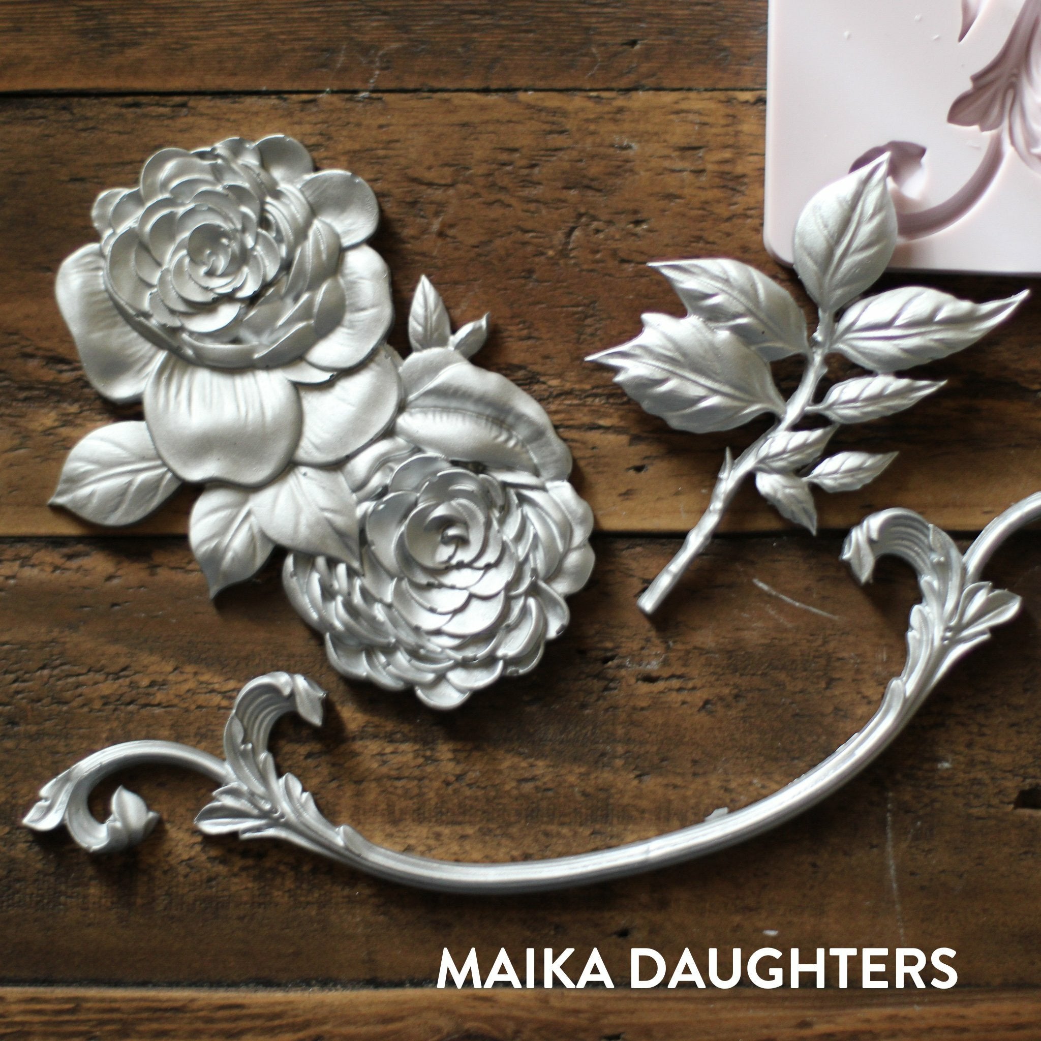 Wooden background with castings of the Victorian rose mold in silver. A white Maika Daughters logo is in the bottom right corner