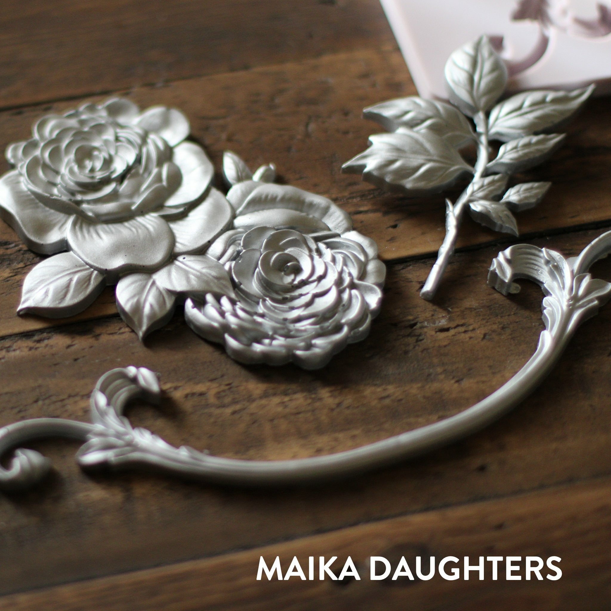 Wooden background with a angled view of the Victorian Rose castings in silver. A white Maika Daughters logo is in the bottom right corner