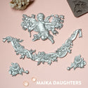 Silver colored castings of a cherub, loose roses, and a rose garland with a ribbon are on a light pink background.