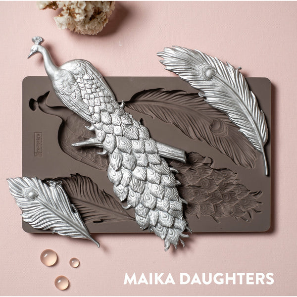 A brown silicone mould and silver colored castings of ReDesign with Prima's Regal Peacock are against a light pink background.
