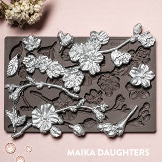 A brown silicone mould and its silver colored castings of cherry blossom sprigs are on a light pink background.