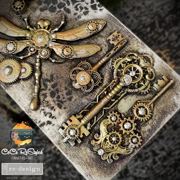 Close-up of a small wood box refurbished by CeCe ReStyled is painted light grey and features ReDesign with Prima's Mechanical Lock & Keys silicone mould castings in bronze color surrounded by antiquing glaze.