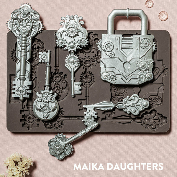 A brown silicone mould and silver colored castings of ReDesign with Prima's Mechanical Lock & Keys are against a light pink background.