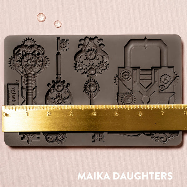 A brown silicone mould of ReDesign with Prima's Mechanical Lock & Keys is against a light pink background. A gold ruler reading 8 inches sits on the mould.