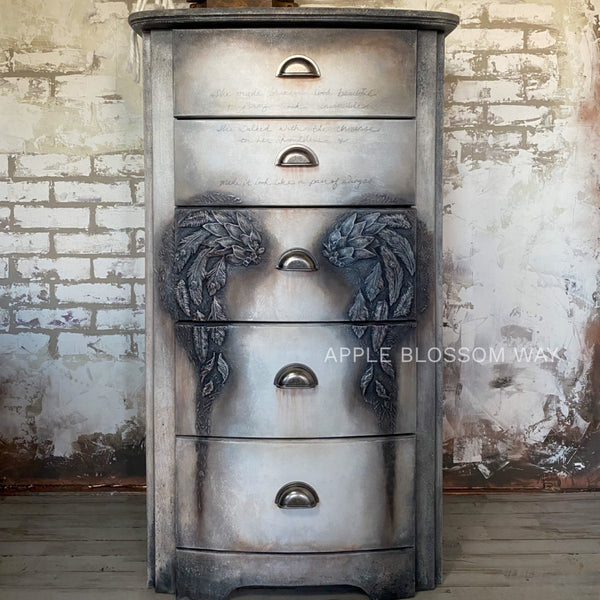 A 5-drawer chest dresser refurbished by Apple Blossom Way is painted a blend of light grey and black and features ReDesign with Prima's Regal Peacock silicone mould on the center drawer.