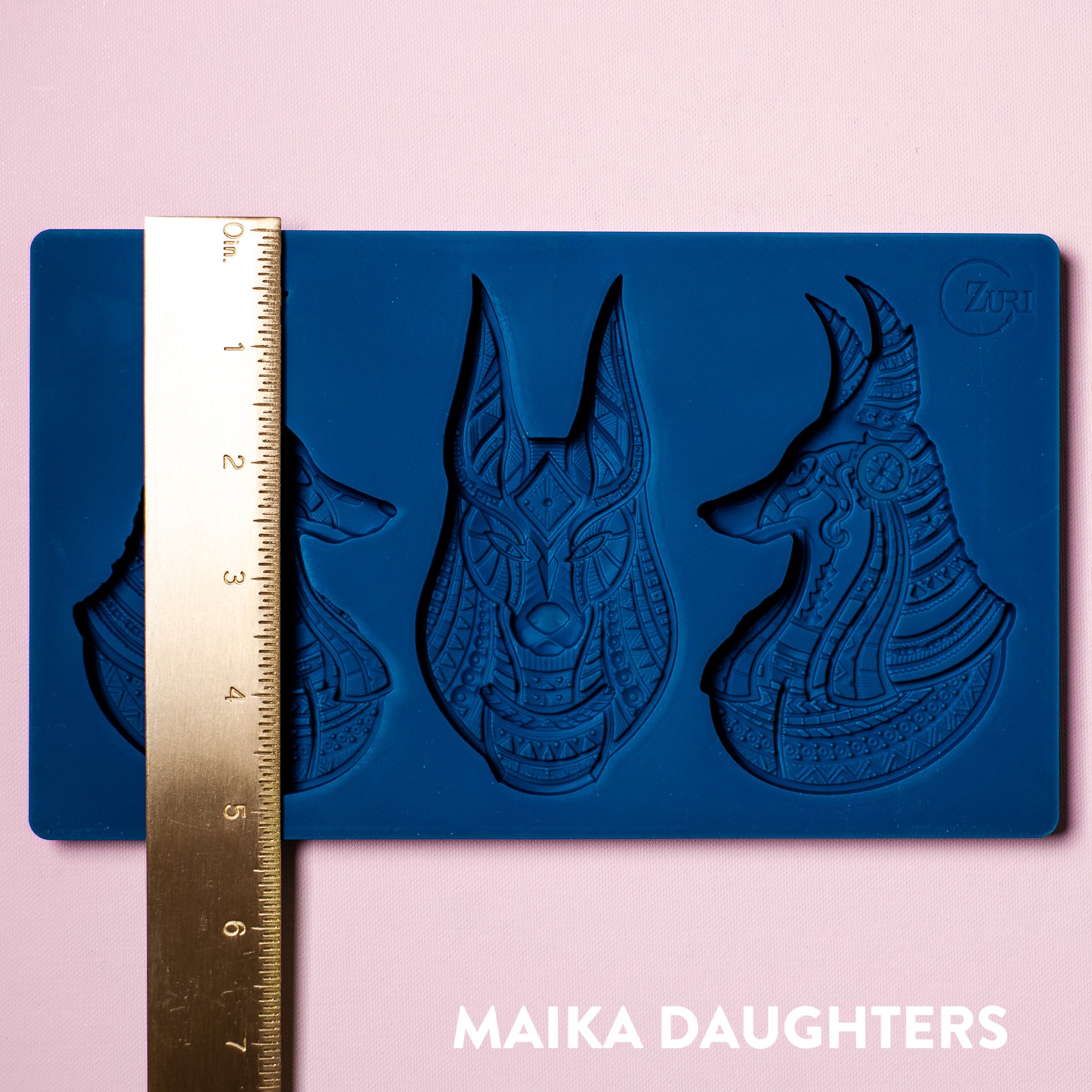 A blue silicone mold of Zuri's Anubis Heads is on a light pink background. A gold ruler showing a measurement of 5.25 inches is on top of the mold.