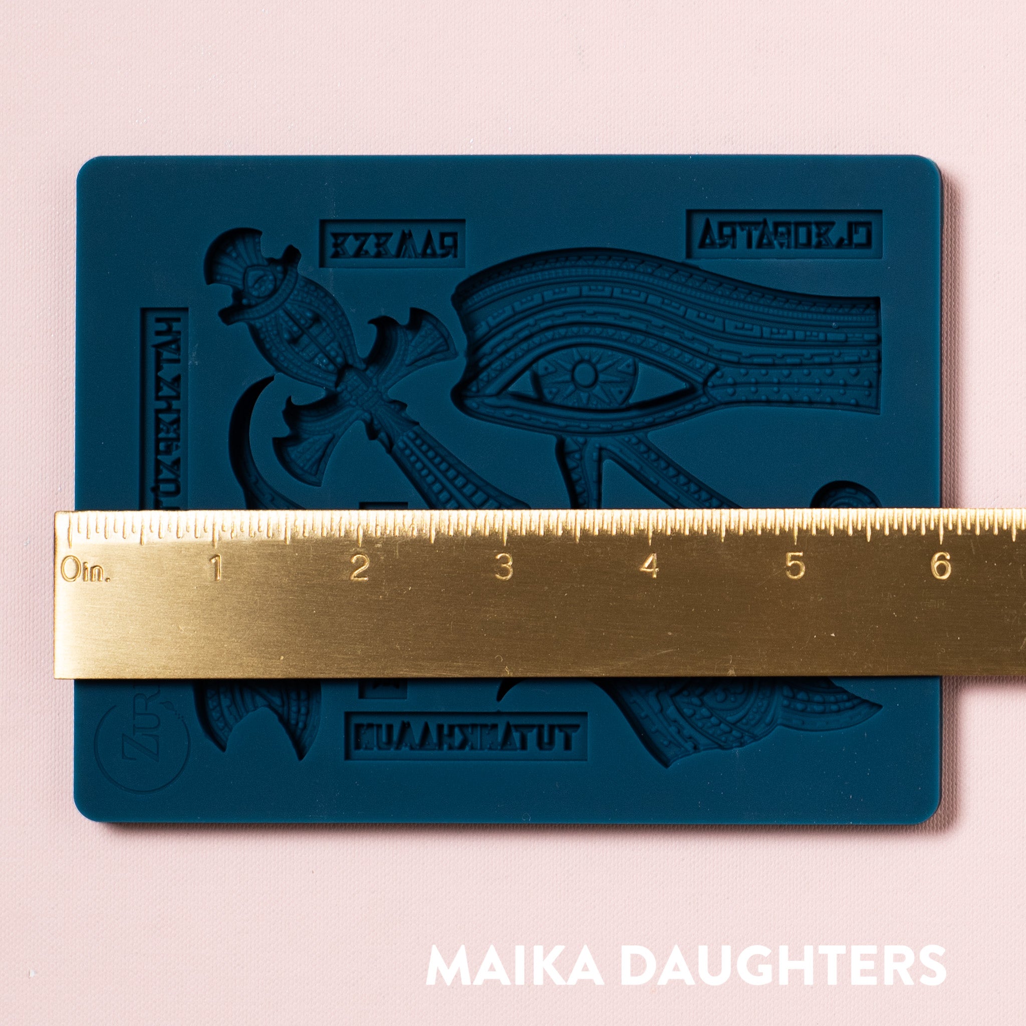 A blue silicone mold of Zuri's Eyptian Motifs is on a light pink background. A gold ruler showing the measurement of 6 inches sits on the mold.