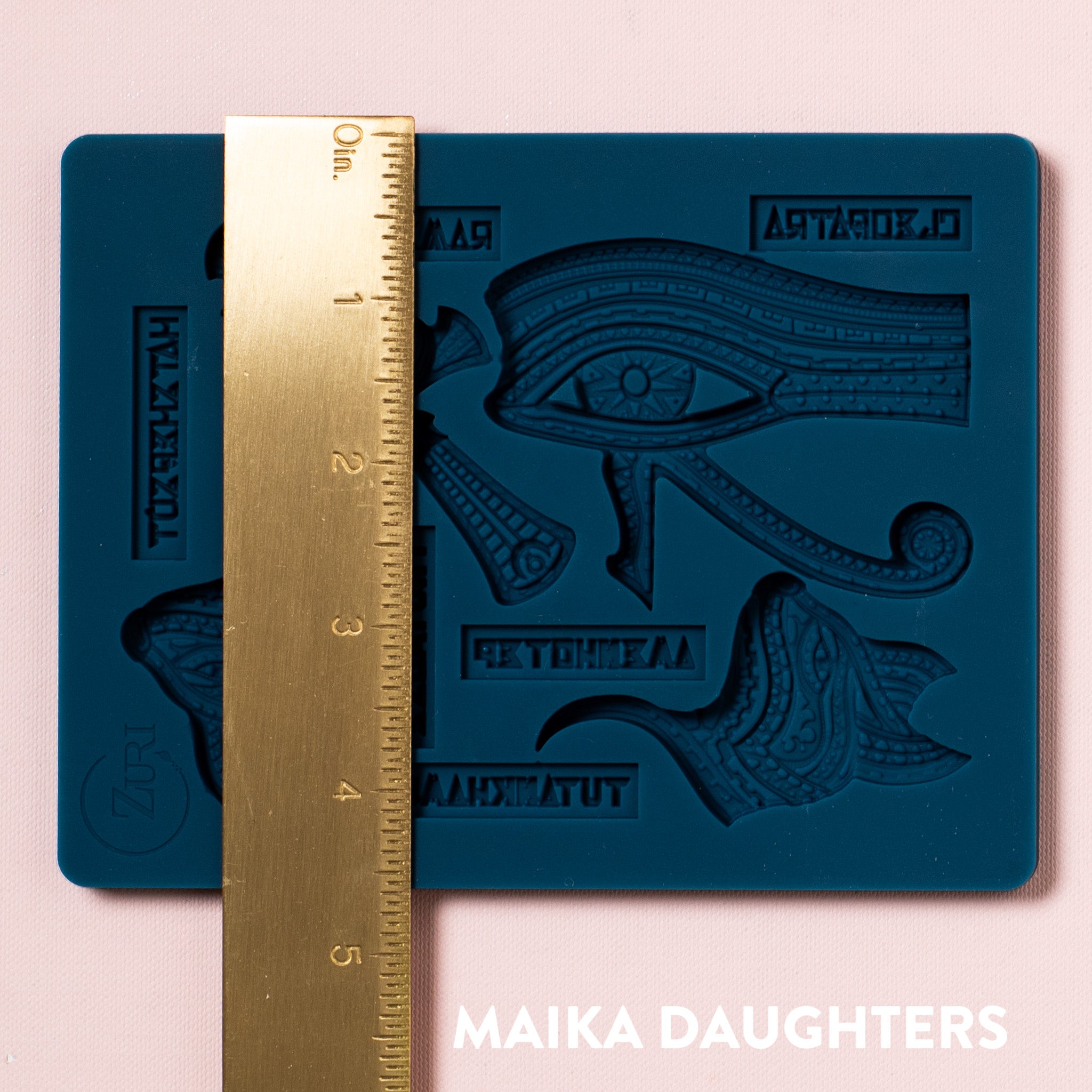 A blue silicone mold of Zuri's Eyptian Motifs is on a light pink background. A gold ruler showing the measurement of 4.5 inches sits on the mold.